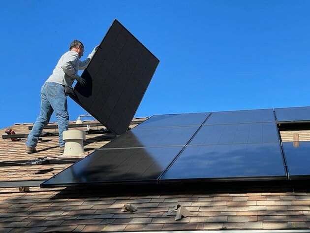  Männliche Person steht auf einem Hausdach und montiert Photovoltaikelemente