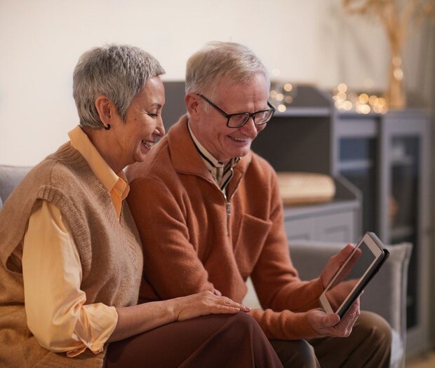  Seniorenpaar sitzt auf dem Sofa und hält ein Tablet in Händen